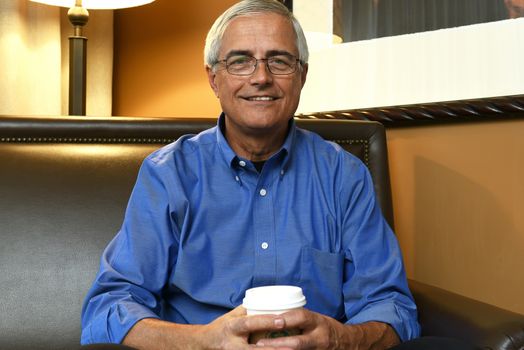 Portrait of a senior businessman seated on the sofa in a hotel room holding a cup of coffee.