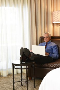 A senior businessman seated on the couch of his hotel room with his feet up while he works on his laptop computer.