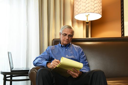 A senior businessman seated on the couch of his hotel room taking notes on a legal pad. 