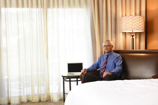 Friendly senior businessman in a hotel room. Man is sitting on the couch next to a table with a laptop computer. Business travel concept.