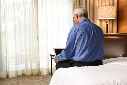 A senior businessman seated on the end of the bed his hotel room while using a tablet computer. The man is seen from behind.