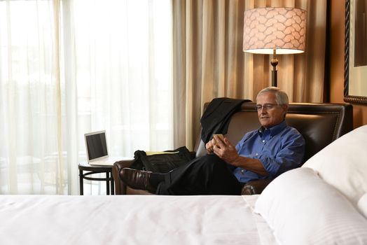 A senior businessman seated on hotel room armchair using his cell phone. Horizontal format with the bed in the foreground 