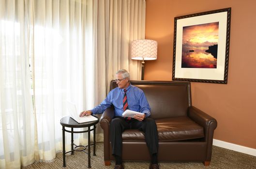 A senior businessman seated on the couch of his hotel room working on his laptop computer.