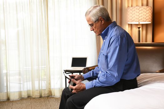 A senior businessman seated on the end of the bed his hotel room while using a tablet computer.