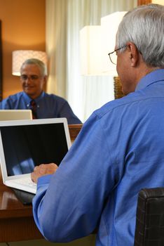 Business Travel Concept: Mature businessman sitting at his hotel room desk working on his laptop computer with his reflection in the mirror.