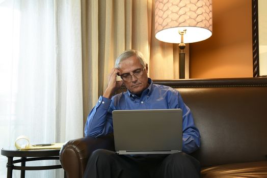 Business Travel Concept: Mature Businessman sitting in his hotel room working on his laptop computer.