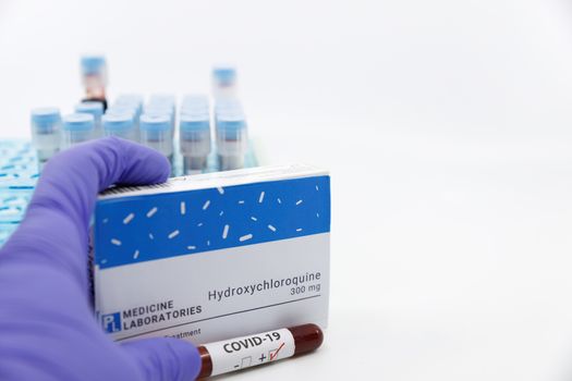 Dubai-UAE-Circa 2020:Doctor showing box of medicine with positive covid-19 test.Concept of Hydroxychloroquine medicine with blood tests tubes on the background.Cure for coronavirus,COVID-19 treatment.