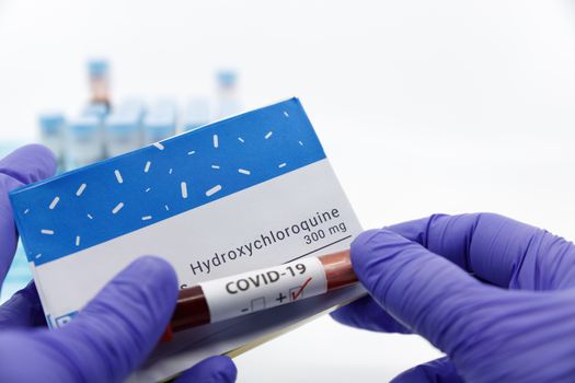 Dubai-UAE-Circa 2020:Doctor showing box of medicine with positive covid-19 test.Concept of Hydroxychloroquine medicine with blood tests tubes on the background.Cure for coronavirus,COVID-19 treatment.