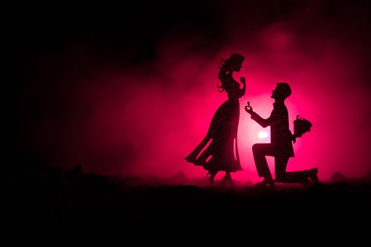 Will you marry me? Silhouette of young man staying on the knee and making proposal for his lovely girl against dark toned background. Valentine greeting card decor. Selective focus