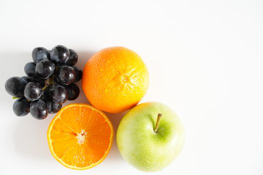 A granny smith green apple, some black grapes and an orange against a plain whit background