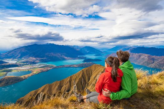 New Zealand hiking couple on mountain top Roys Peak relaxing sitting enjoying active outdoor lifestyle looking at view of amazing New Zealand nature landscape, near Wanaka, Otago, South Island.