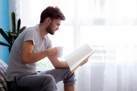 Young man reading book and drinking coffee on sofa in the living room