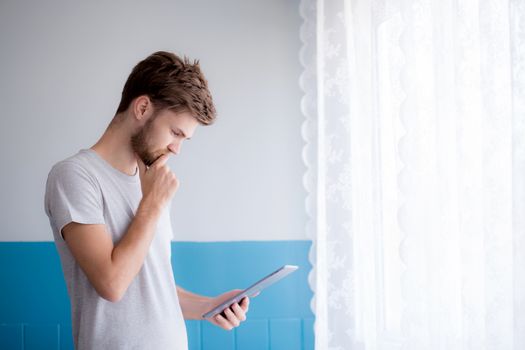 Closeup view of pensive bearded man using tablet while standing near the window in his modern apartment.Concept young business people working at home.