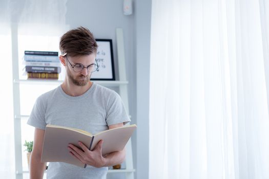 Handsome guy in eyeglasses is reading book preparing exam with standing at the living room - education concept.