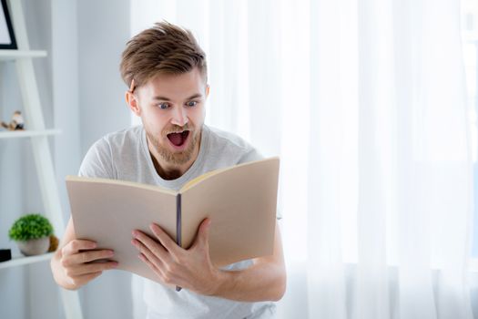 Handsome guy in eyeglasses is reading book preparing exam with standing at the living room, education concept