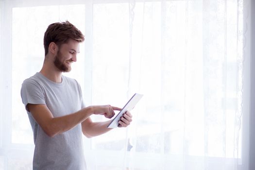 Closeup view of pensive bearded man using tablet while standing near the window in his modern apartment - Concept young business people working at home.