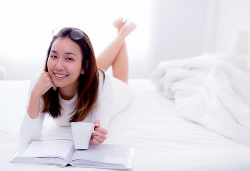 close up of a charming woman lying on bed reading a book and holding coffee cup looking into camera in bedroom.