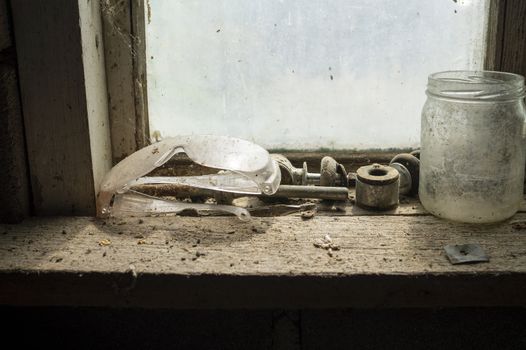 Safety goggles with old tools on a dusty wooden workbench with empty glass jar in front of a window
