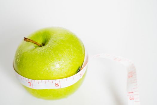 A granny smith green apple with a measuring tape wrapped around it against a plain white background