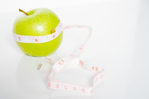 A granny smith green apple with a measuring tape wrapped around it against a plain white background