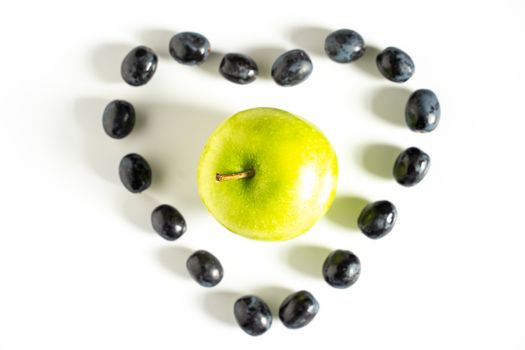 A green granny smith apple in the center of a heart shape made up of some black grapes