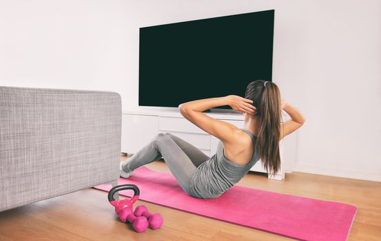 Home fitness woman doing strength training abs situps bodyweight floor exercises watching online livestream workout, web videos casted on smart tv in living room of house or apartment.