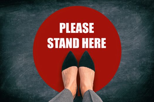 COVID-19 Coronavirus message asking supermarket customer to stand in space. Top view of feet standing in red circle with text in public space practicing social distancing. Blackboard background.