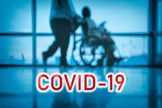 COVID-19 billboard red text on blue medical hospital background with doctor walking with disabled patient in wheelchair.