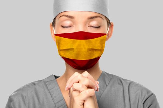 COVID outbreak in Spain Asian doctor with spanish flag print on surgical mask praying for help. Young woman medical nurse praying. Coronavirus pandemic in Spain.