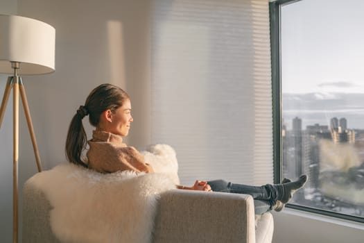 Staying home Asian woman relaxing pensive looking out the window sitting in living room sofa enjoying free time to think and practice mindfulness, indoors during coronavirus outbreak.