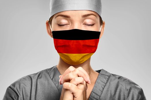 German doctor praying for help with Germany flag on surgical mask. Young woman medical needing PPE help to fight COVID-19 Coronavirus .