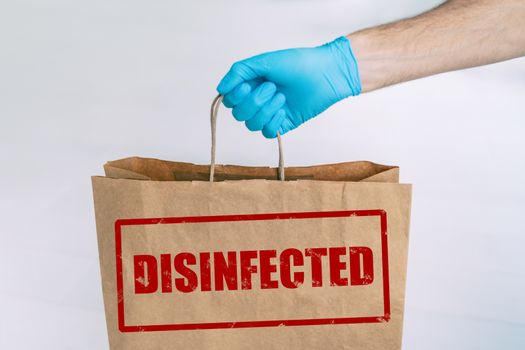 Delivery bag to home from online grocery shopping with stamp of safety DISIFINFECTED written on the paper side. Man holding groceries with medical blue glove.