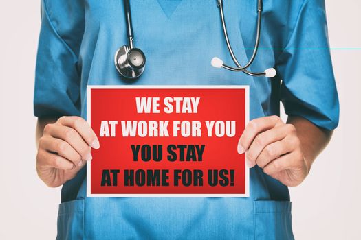 COVID-19 Coronavirus doctor holding sign for viral social media post showing: We Stay at Work for You, Please you stay at home for us! on card. Nurse with blue srubs isolated on white background.