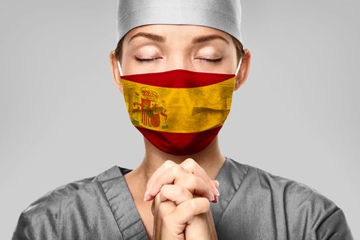 COVID-19 Spain flag print on mask of Asian spanish medical doctor praying in hope for help.