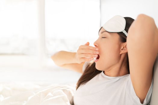 Waking up early morning at sunrise happy woman yawning stretching in bed. Sleeping at home woman waking up in morning sunlight stretching happy after good night sleep feeling rested wearing eye mask.