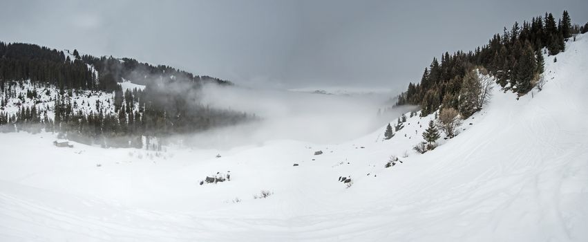 Panoramic view of a snow covered mountain range looking down valley with low clouds and an overcast sky