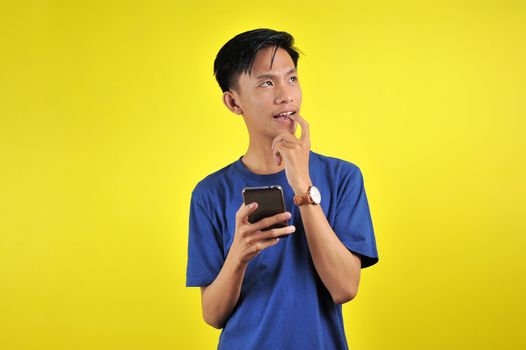 Happy young Asian man smiling using smartphone looking at blank area, isolated on yellow background