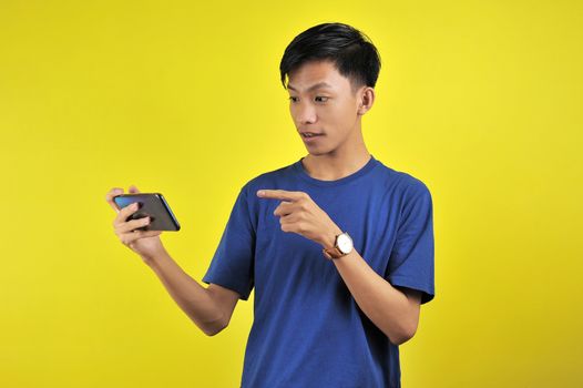 Shocked face of Asian man in white shirt looking at phone screen on yellow background.