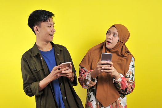 Photo of positive excited people man and woman screaming and looking at each other while both using mobile phones isolated over yellow background