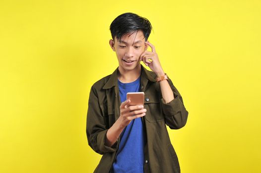 Portrait of smart person Young Asian man doing thinking gerture look at smartphone display, isolated on yellow