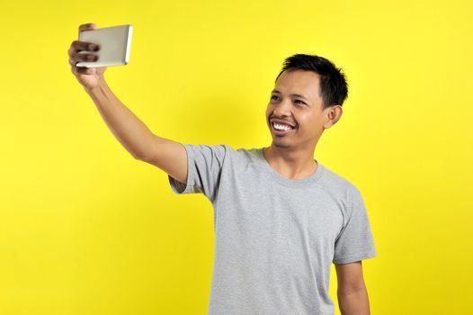 Portrait of cool cheerful Asian man having video-call with lover holding smart phone in hand shooting selfie on front camera, isolated on yellow background