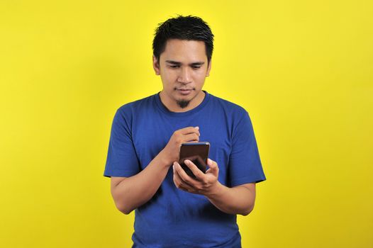 STRAIGHT FACE of Asian man in white shirt looking at phone screen on yellow background.