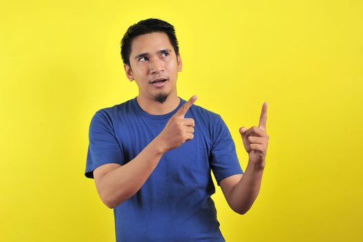 Friendly-looking lively pleasant Asian male with white t-shirt smiling delighted look at copy space joyful. Self-assured pointing, on yellow background