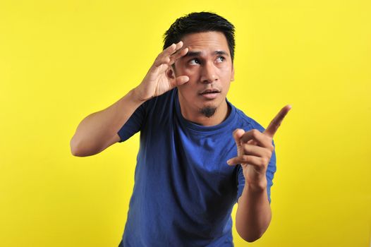 Young Asian man open his eyes and pointing the blank area, isolated on yellow background