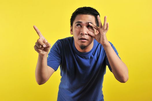 Young Asian man open his eyes and pointing the blank area, isolated on yellow background