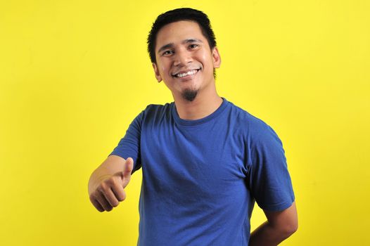 Portrait of young Asian man laughing with showing thumbs up at camera, isolated on yellow