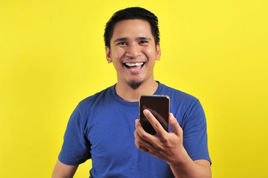 Happy young good looking Asian man smiling using cellphone, isolated on yellow background