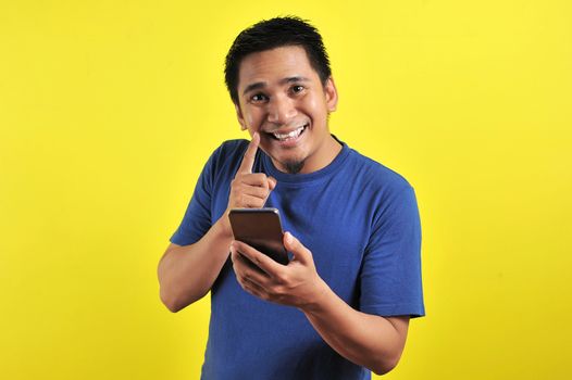 Happy young good looking Asian man smiling using cellphone, isolated on yellow background