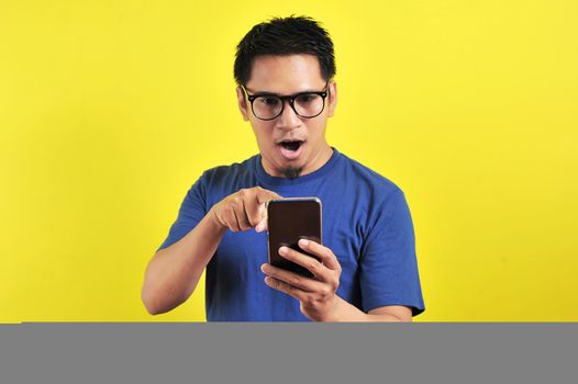 Shocked face of Asian man in blue shirt looking at phone screen on yellow background.