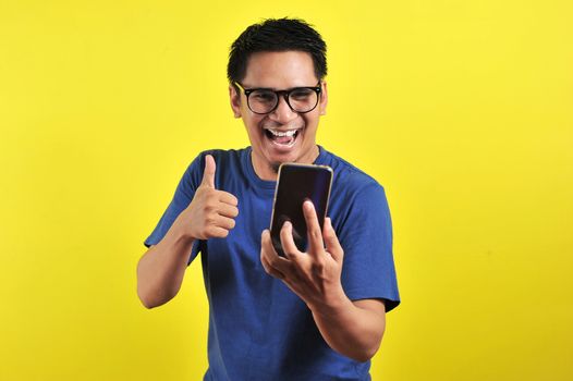 Shocked face of Asian man in blue shirt looking at phone screen on yellow background.